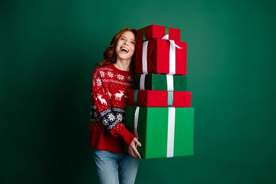 Happy young woman in red Christmas sweater carrying a tall stack of wrapped gifts against a green background