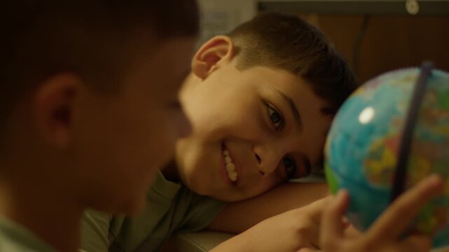 Two young boys smile while holding a globe together. One boy leans in, eyes wide with curiosity. Their hands gently cradle the colorful map. Warm indoor lighting adds cozy charm