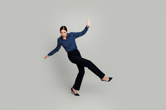 Businesswoman losing balance in high heels on gray background, surprised woman mid fall wearing polka dot blouse and black trousers
