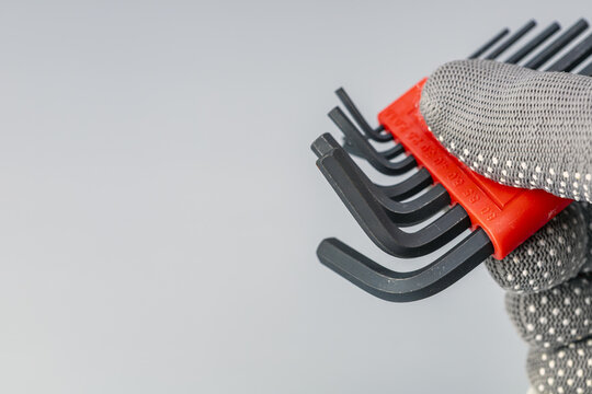 Detailed close-up view of a worker hand wearing a gray dotted protective fabric glove holding a complete set of black L-shaped steel hex allen key wrenches arranged by size in a bright red plastic