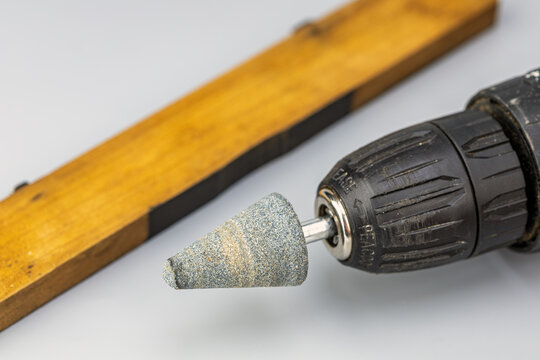 Detailed macro close up of a gray conical abrasive mounted grinding stone bit with metal shank secured in the keyless chuck of a black cordless power drill, with a blurred wooden plank on a white