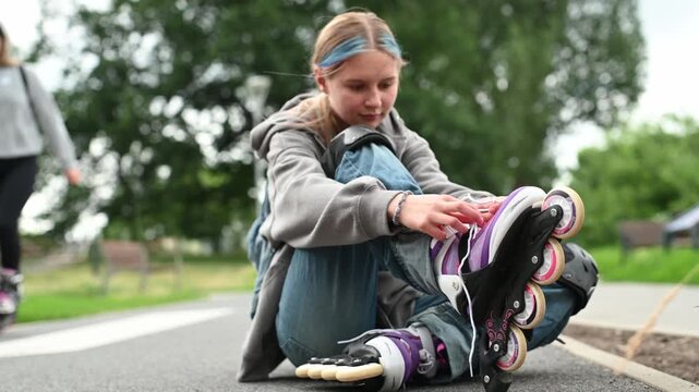 Young girl sits on paved ground, adjusting her purple and white rollerblade during daytime.