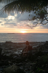 Silhouette of a woman sitting on beach rocks at sunset under palm leaves in Puntarenas, Costa Rica © Julin