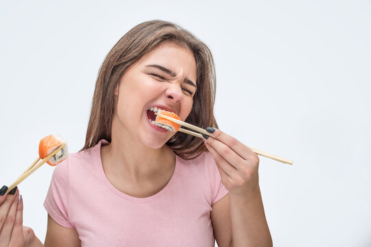 Young woman devour pieces of sushi. She bite piece. Model hold them with chopsticks. Isolated on grey background.
