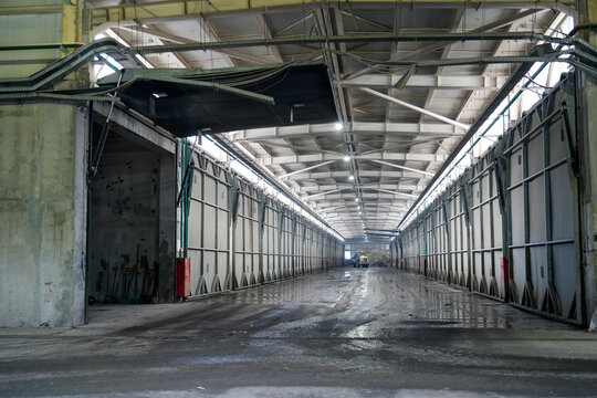 A long industrial waste facility corridor with metal wall panels, exposed roof trusses and a wet concrete floor, showing the interior of a large waste processing building.