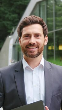 Handsome bearded businessman with laptop standing in front of a modern office building, looking at the camera with confidence. Successful entrepreneur and corporate executive in an urban business