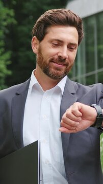 Successful businessman in a formal suit walking through a modern urban park near a contemporary glass office building. Smiling professional checking the time on his wristwatch while heading to a