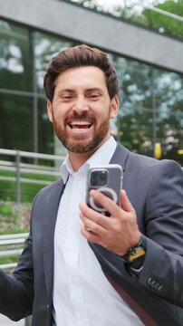 Happy businessman checking his smartphone and smiling while standing in a city park near a modern office building, enjoying positive news and successful business communication. Slow motion