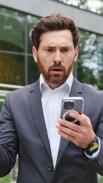 Disappointed businessman checking email on his smartphone in a city park near a modern office building, reacting to bad news with frustration, stress, and negative emotions in a professional business