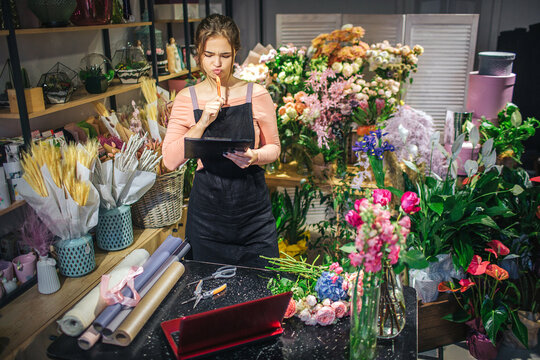 Serious and concentrated young florist hold black planchette. She think and look at it. Floris is surround with flowers and plants. Paper rolls and red laptop are on table.