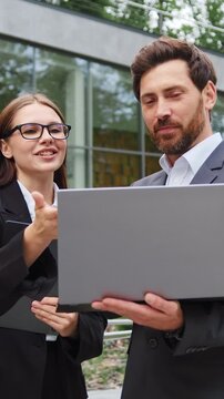 Businessman and businesswoman smiling and discussing work while looking at a laptop screen outdoors. Teamwork, business communication, collaboration, and modern work lifestyle. Slow motion