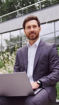 Handsome bearded businessman in a suit sitting on a bench in a city park, working on a laptop and thoughtfully looking into away. Slow motion
