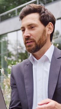 Businessman sitting outdoors in a city park, working on a laptop and having a professional video call while looking at the screen and greeting a business partner. Slow motion