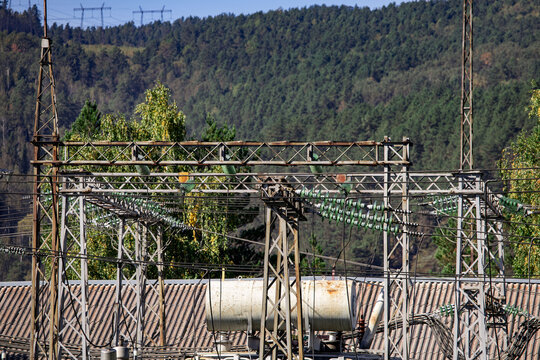 Complex network of electrical power lines and metal transmission towers situated in an industrial area, set against a backdrop of dense green forest and hills. Various cables and support structures cr