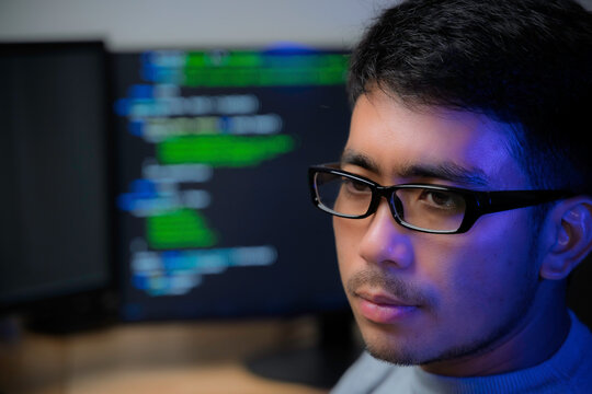 Computer programmer works at a multi monitor workstation in a dark office, reviewing programming code for software development, technical analysis, and system monitoring.