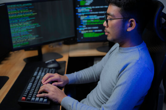 Computer programmer works at a multi monitor workstation in a dark office, reviewing programming code for software development, technical analysis, and system monitoring.