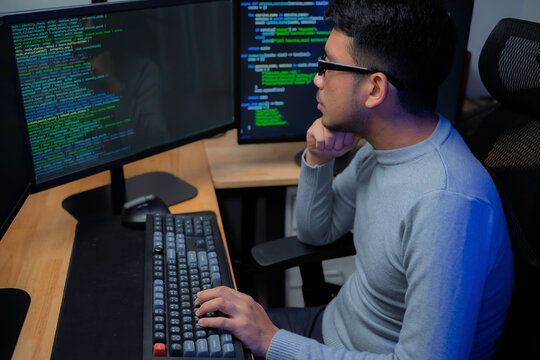 Computer programmer works at a multi monitor workstation in a dark office, reviewing programming code for software development, technical analysis, and system monitoring.