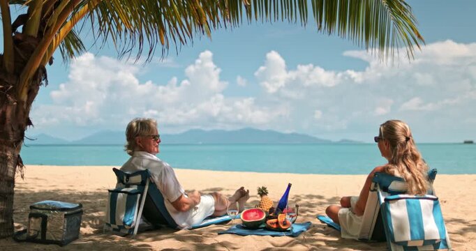 Senior couple talking and relaxing on beach chairs under a palm tree, sharing a picnic with fresh fruit and drinks while looking out at the turquoise ocean on a beautiful sunny day in Samui, Thailand
