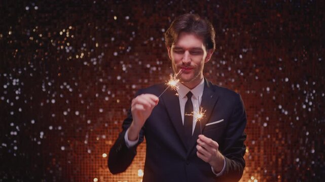 Smiling young man in an elegant suit holding sparklers with glowing sparks and abstract bokeh lights in the background, celebrating New Year, Christmas, birthday or festive occasion. Handsome male
