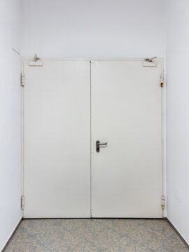 closed double-leaf white metal door, typical of emergency exits, fire-rated or utility doors, set against clean white walls and a grey tiled floor in a modern, minimalist building corridor.