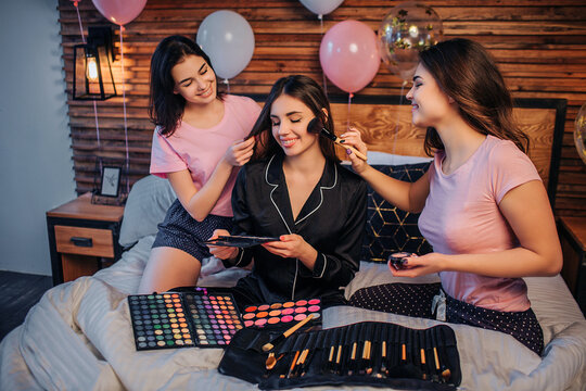 Three happy young women sit on bed in festive room. Two girls do haircare and makeup for third one. There are brushes and shadown on bed. Model in black pajamas hold one set of shadows in hands.
