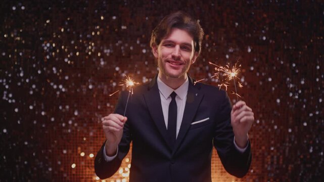 Smiling young man in an elegant suit holding sparklers with glowing sparks and abstract bokeh lights in the background, celebrating New Year, Christmas, birthday or festive occasion. Handsome male