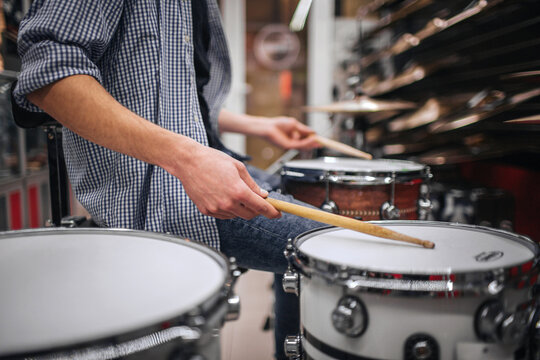 Picture of young bearded man playing on drus. He touches them with special sticks. Many cymbals are above on shelfs. Guy is alone in room.
