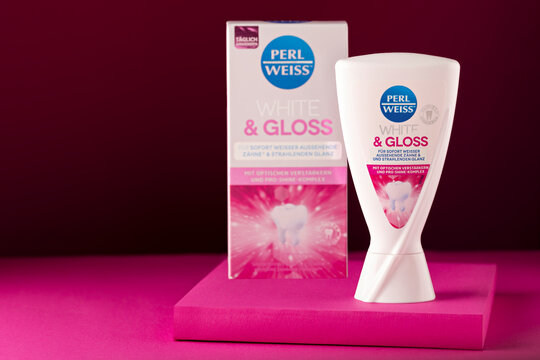 Perl Weiss White & Gloss whitening toothpaste displayed in front of its matching retail carton on a pink platform against a burgundy background. Selective focus emphasizes the toothpaste container, wi