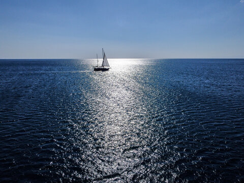 Sailboat cruising on sparkling open water under a bright, sunny sky, aerial drone view. White yacht sailing smoothly across the deep blue sea. Boat navigating calm waters in summer. Sailing vessel 