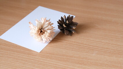 Empty white greeting card with dried flower and pine cone on a natural wooden desk with copy space. Minimalist autumn or winter stationery concept. © Michel F