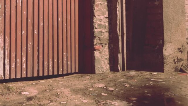 Sunlight bathes an old brick wall in soft hues, while a rust colored gate stands partially open. Dusty ground and scattered stones create a rustic charm in this peaceful alleyway.