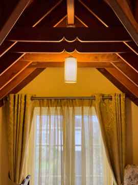 warm wooden cottage interior with a-frame ceiling, hanging cylindrical lamp and window with sheer curtains, cozy rustic home design concept with copy space