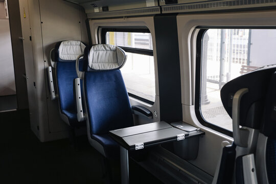 Empty blue passenger train seats beside windows. White fabric headrest covers near black folding table. Railway carriage interior background. Gray panels near platform outside. Quiet travel seating.