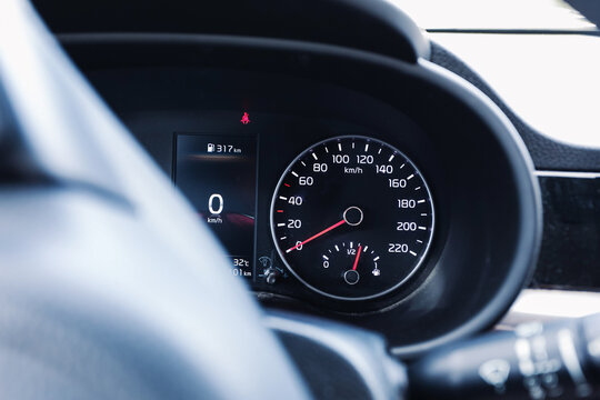 Car dashboard with analog speedometer at zero. Digital speed display with fuel range indicator. Red seat belt warning light background. Black instrument cluster by steering wheel. Interior details.