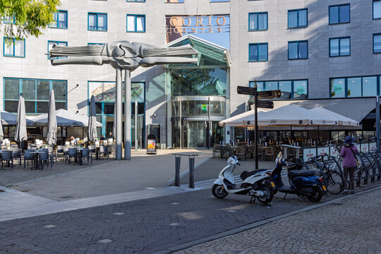 Heerlen, Netherlands - 04 October 2026: Entrance of the Corio Center shopping mall featuring modern artwork sculptures, outdoor cafe dining terraces, and parked motor scooters.