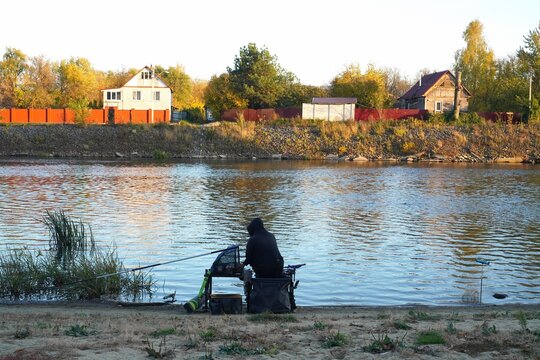 Fisherman in hoodie sitting on angler platform with rod fishing on autumn river at sunset. Village houses and golden trees on opposite bank, seasonal fall angling with modern platform gear.