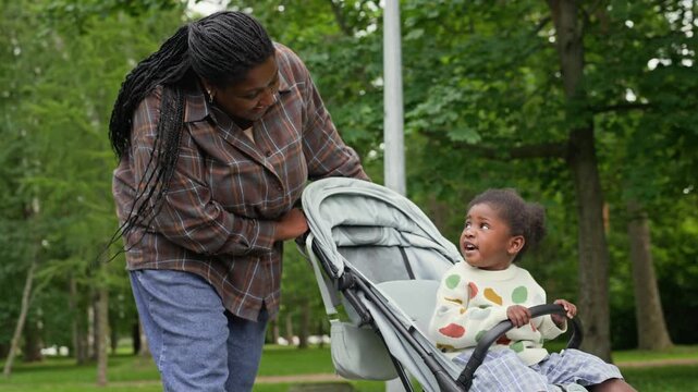 African American mother pushing stroller with toddler daughter while walking in summer park. Footage suitable for parenting campaigns, maternal health promotions and family lifestyle advertising