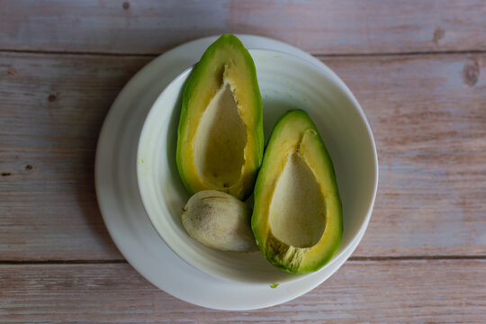 Overhead view of a ripe avocado (Persea americana) cut lengthwise into two halves with the large seed removed and placed beside the fruit in a white ceramic bowl. Creamy yellow green flesh and natural