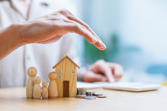 Person hands protecting miniature wooden house and family figurines with stacks of coins on table, home insurance and financial planning concept