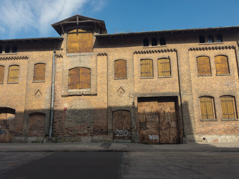 Exterior of an old brick industrial building with boarded-up windows, weathered wooden shutters and graffiti-covered doors. The deteriorating facade reflects urban decay and industrial architecture