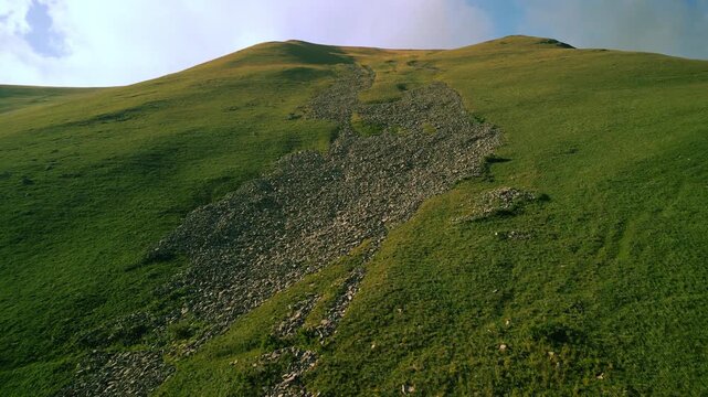 Aerial Drone Flight Climbing Up Green Mountain Slope Past Rocky Scree Field Toward Blue Sky and White Clouds in Armenia