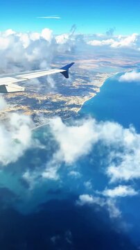 Aerial view from an airplane window of a wing with a blue winglet over a sunny coastline, resort town and blue sea with clouds, travel and vacation concept, grainy film texture