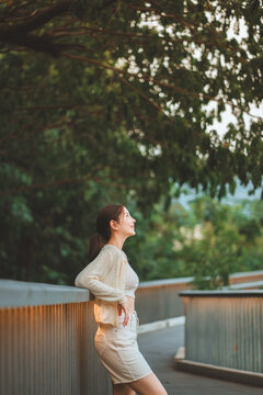 Happy young Asian woman relaxing outdoors while leaning against railing in peaceful green park, smiling and enjoying fresh air, warm sunlight, nature, emotional wellbeing,