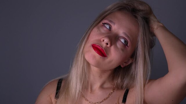 Young blonde woman with red lips adjusts her hair with a haughty diva expression, showing confidence, glamour and bold feminine attitude in a close beauty portrait