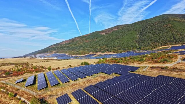 Rows of solar panels stretch across a hillside in a rural area. Bright sunlight shines on the panels, which collect energy. The sky is clear with some clouds and trails.