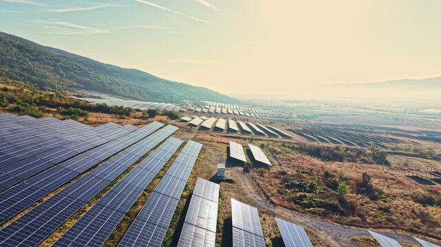 A vast field of solar panels stretches across a valley. The sun shines brightly in the sky. The background consists of green hills.