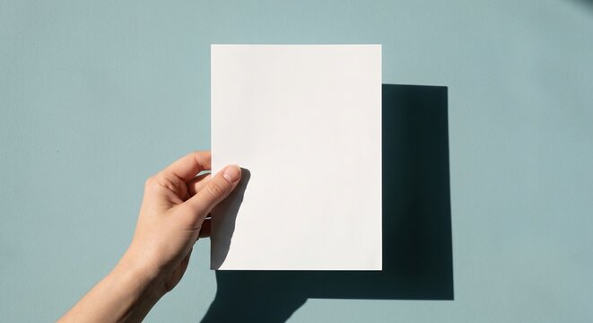 A hand holds a blank matte white flyer against a light teal-blue wall with angular shadows.