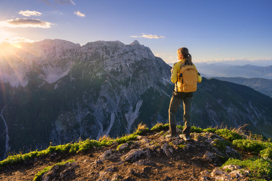 Woman hiker with yellow backpack standing on rocky mountain summit in Slovenia. Alpine autumn landscape with panoramic view of peaks, sky, clouds, green hills, sun flare and evening sunset light