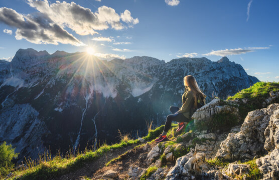 Woman hiker with backpack sitting on rocky hill and looking at mountains at sunset in Slovenia. Alpine autumn landscape with mountain peaks, sky, clouds, sunbeams, sun flare and evening sunlight
