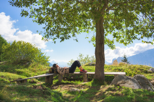 Woman hiker lying down on wooden bench resting under green tree in Slovenia. Alpine landscape with girl, mountains, blue sky, clouds, green grass meadow, backpack and sunny outdoor view in summer